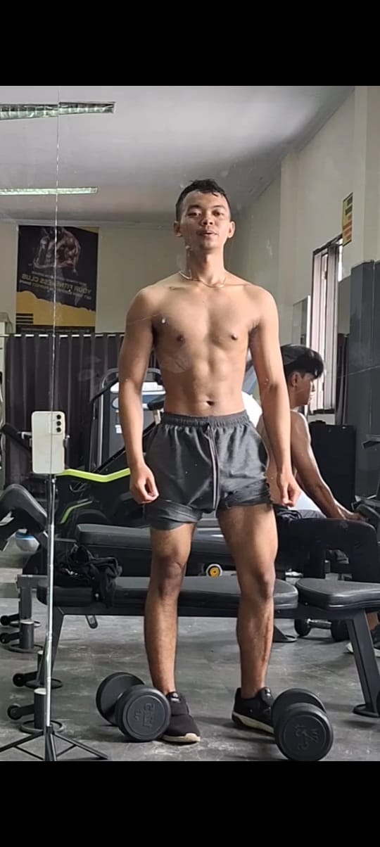 Foto Dicky Darmansyah sedang berdiri tegak di depan alat pengangkat berat, tanpa mengenakan atasan dan memakai celana pendek. Ia tampak menatap ke arah cermin dengan ekspresi fokus.